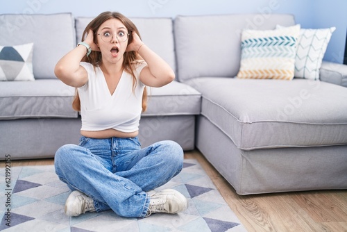 Young caucasian woman sitting on the floor at the living room crazy and scared with hands on head, afraid and surprised of shock with open mouth