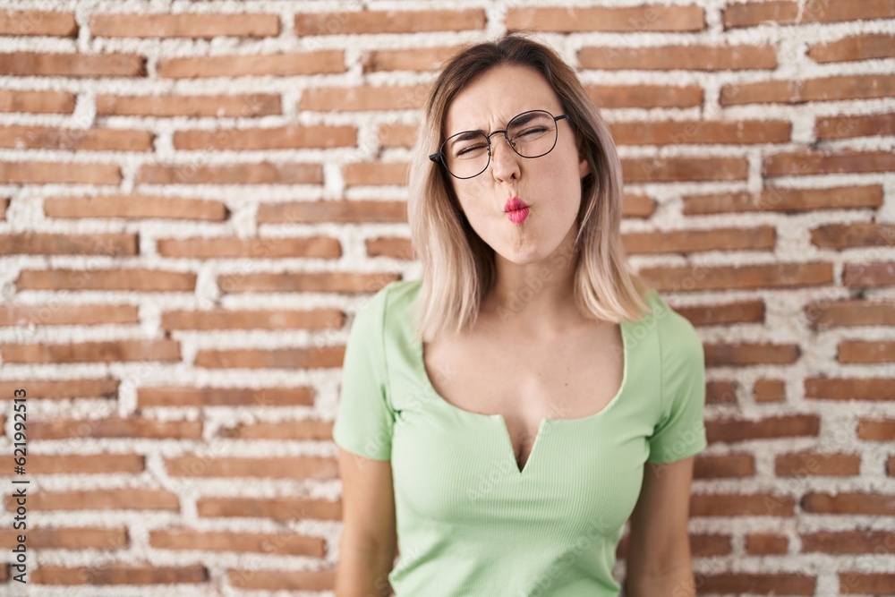 Young beautiful woman standing over bricks wall making fish face with lips, crazy and comical gesture. funny expression.