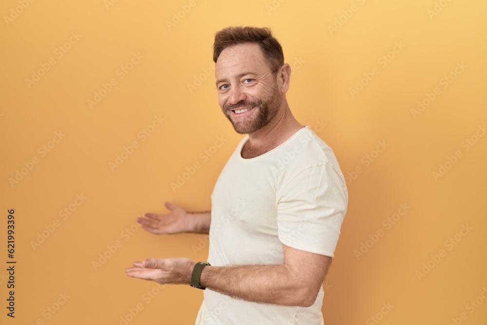 Middle age man with beard standing over yellow background inviting to enter smiling natural with open hand