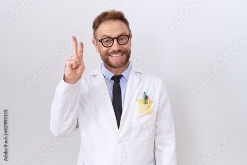 Middle age doctor man with beard wearing white coat showing and pointing up with fingers number two while smiling confident and happy.