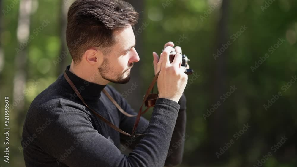 Side view portrait of smiling Caucasian young man taking photos of nature with camera standing in sunshine in summer forest. Happy confident tourist enjoying leisure on vacations Side view portrait of smiling Caucasian young man taking photos of nature with camera standing in sunshine in summer forest. Happy confident tourist enjoying leisure on vacations