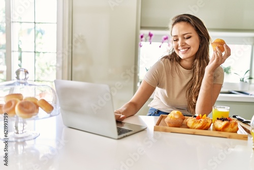 Young beautiful hispanic woman having breakfast using laptop at the kitchen