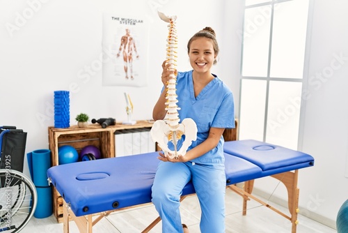 Young beautiful hispanic woman physiotherapist smiling confident holding anatomical model of spinal column at rehab clinic