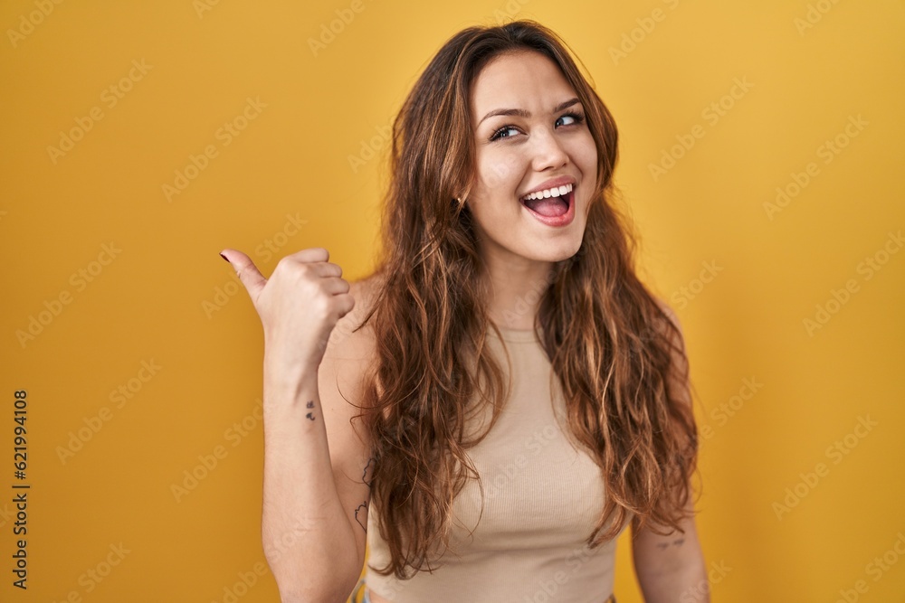 Young hispanic woman standing over yellow background smiling with happy face looking and pointing to the side with thumb up.
