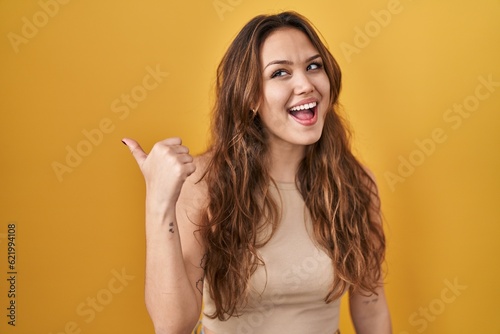 Young hispanic woman standing over yellow background smiling with happy face looking and pointing to the side with thumb up.