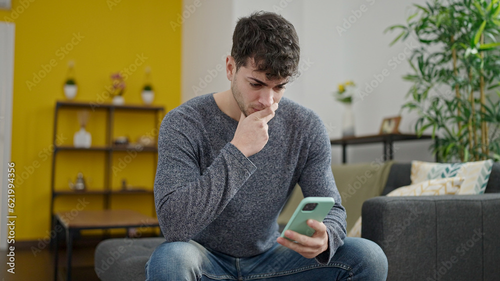 Young hispanic man using smartphone worried sitting on the sofa at home