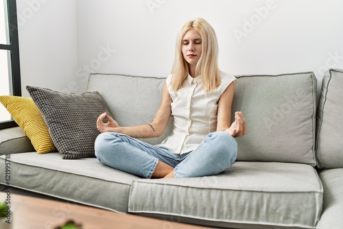 Young blonde woman doing yoga exercise sitting on sofa at home