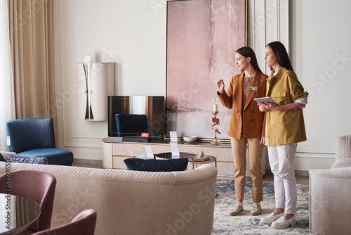 Two young brunette women in smart casualwear discussing features of new pieces of furniture while standing in one of store departments