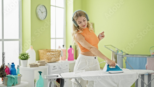 Young blonde woman listening to music ironing clothes dancing at laundry room