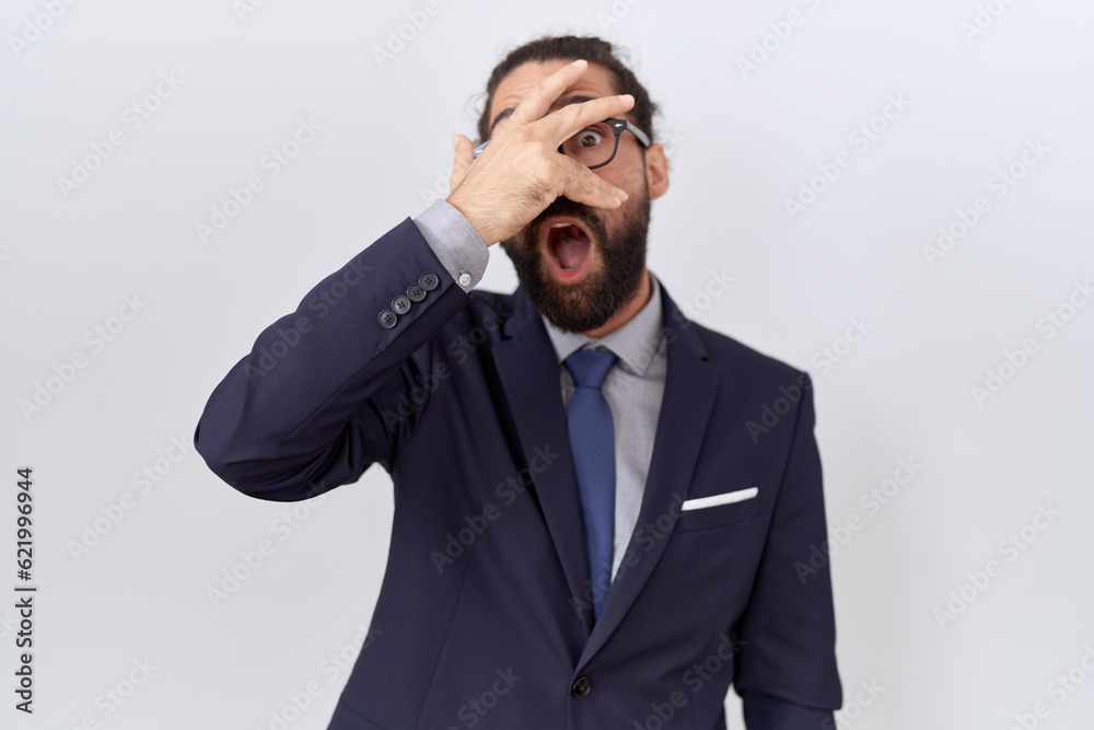 Hispanic man with beard wearing suit and tie peeking in shock covering face and eyes with hand, looking through fingers with embarrassed expression. Hispanic man with beard wearing suit and tie peeking in shock covering face and eyes with hand, looking through fingers with embarrassed expression.