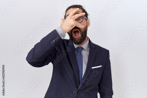 Hispanic man with beard wearing suit and tie peeking in shock covering face and eyes with hand, looking through fingers with embarrassed expression. © Krakenimages.com