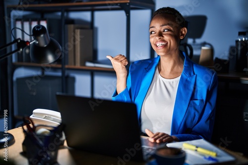 Beautiful african american woman working at the office at night smiling with happy face looking and pointing to the side with thumb up.