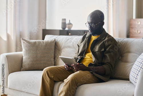 Young African American male freelancer with tablet sitting on soft comfortable couch in living room and looking through online data © pressmaster