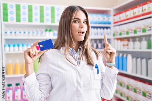 Hispanic young woman working at pharmacy drugstore holding credit card surprised with an idea or question pointing finger with happy face, number one
