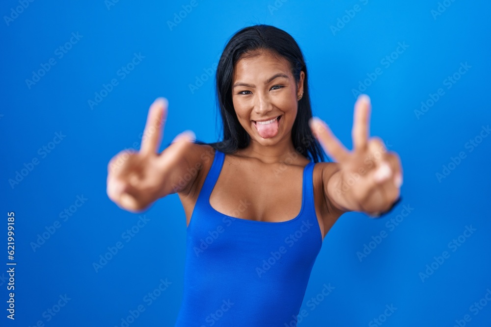 Hispanic woman standing over blue background smiling with tongue out showing fingers of both hands doing victory sign. number two. Hispanic woman standing over blue background smiling with tongue out showing fingers of both hands doing victory sign. number two.