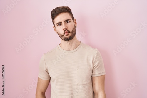 Hispanic man with beard standing over pink background making fish face with lips, crazy and comical gesture. funny expression.