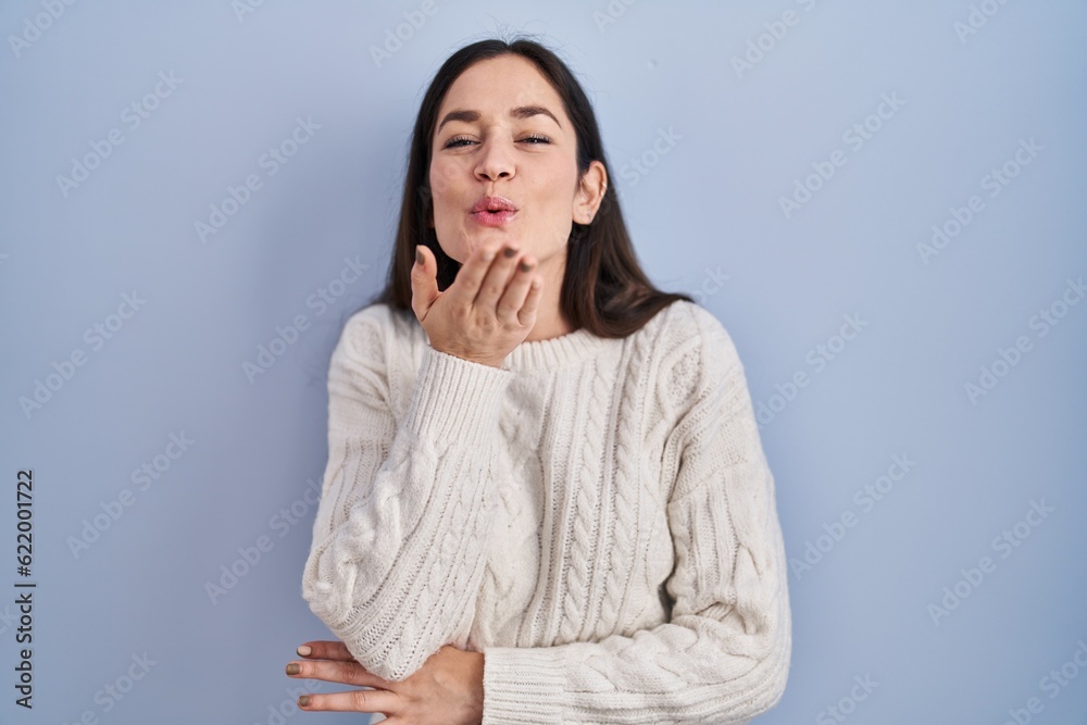 Young brunette woman standing over blue background looking at the camera blowing a kiss with hand on air being lovely and sexy. love expression. Young brunette woman standing over blue background looking at the camera blowing a kiss with hand on air being lovely and sexy. love expression.
