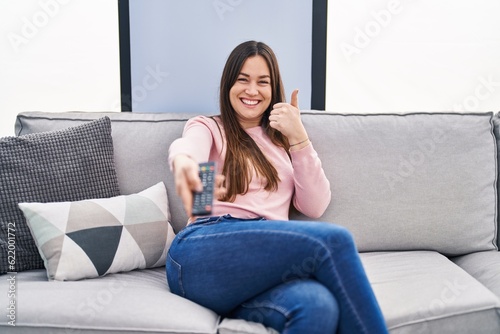 Young brunette woman holding television remote control smiling happy and positive  thumb up doing excellent and approval sign