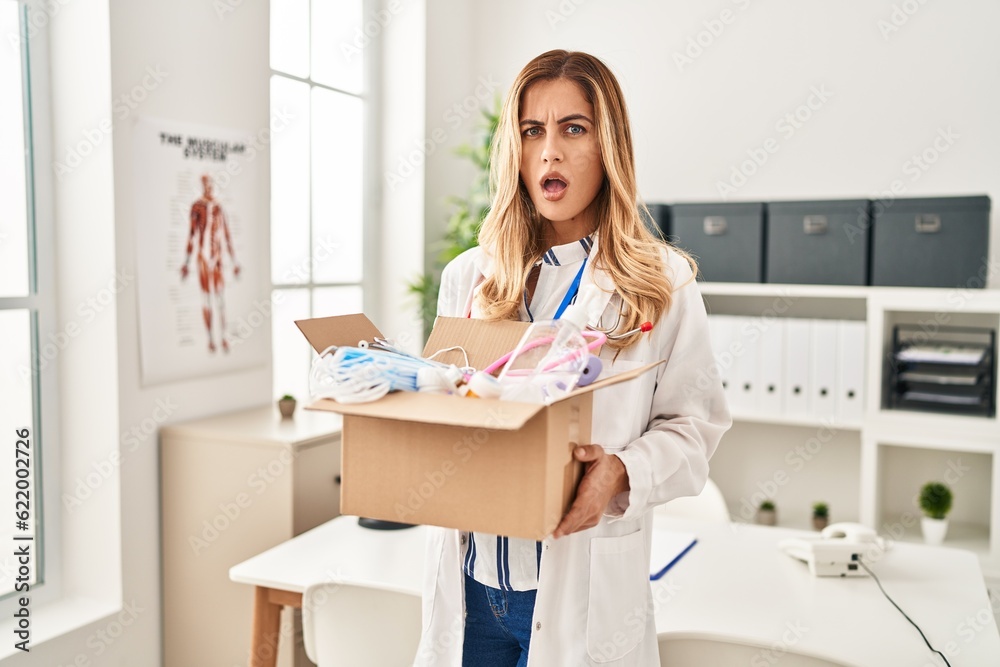Young blonde doctor woman holding box with medical safety items clueless and confused expression. doubt concept.