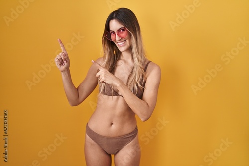 Young hispanic woman wearing bikini over yellow background smiling and looking at the camera pointing with two hands and fingers to the side.