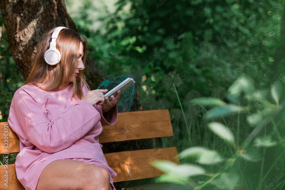Naklejka premium A young beautiful woman listening to music with headphones with her eyes closed on a park bench. Peace, harmony, relaxation and relaxation.