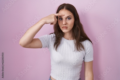 Young hispanic girl standing over pink background pointing unhappy to pimple on forehead  ugly infection of blackhead. acne and skin problem