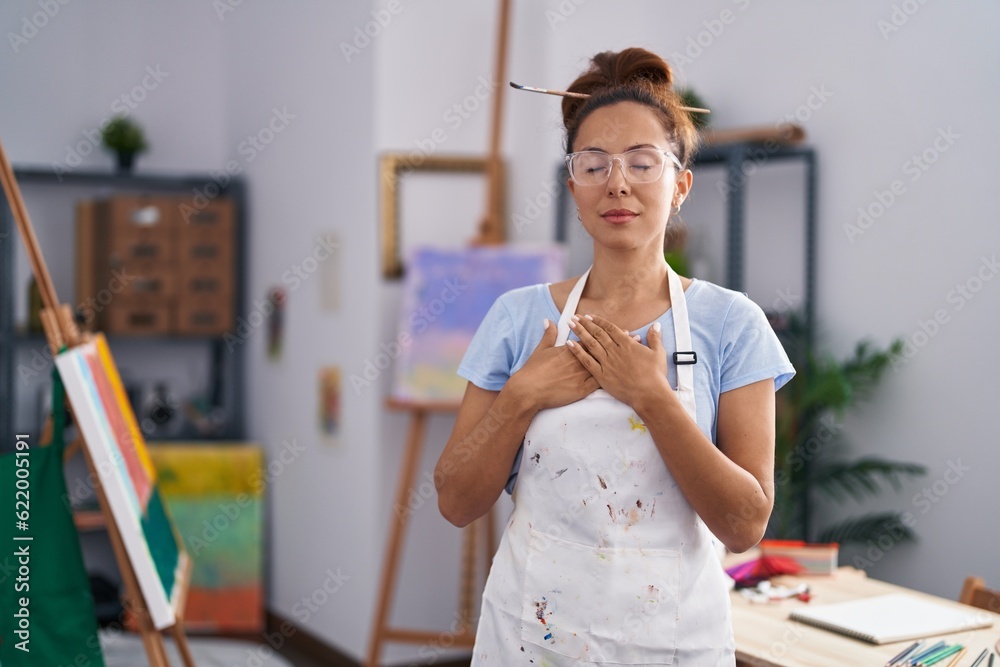 Brunette woman painting at art studio smiling with hands on chest with closed eyes and grateful gesture on face. health concept. Brunette woman painting at art studio smiling with hands on chest with closed eyes and grateful gesture on face. health concept.