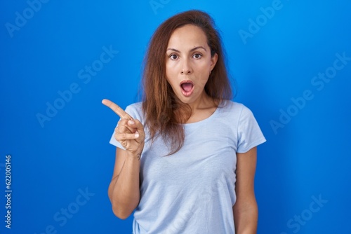 Brunette woman standing over blue background surprised pointing with finger to the side, open mouth amazed expression.