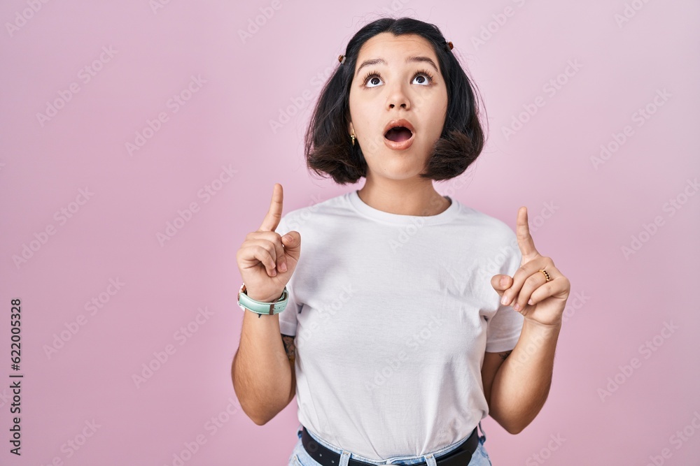 Young hispanic woman wearing casual white t shirt over pink background amazed and surprised looking up and pointing with fingers and raised arms. Young hispanic woman wearing casual white t shirt over pink background amazed and surprised looking up and pointing with fingers and raised arms.