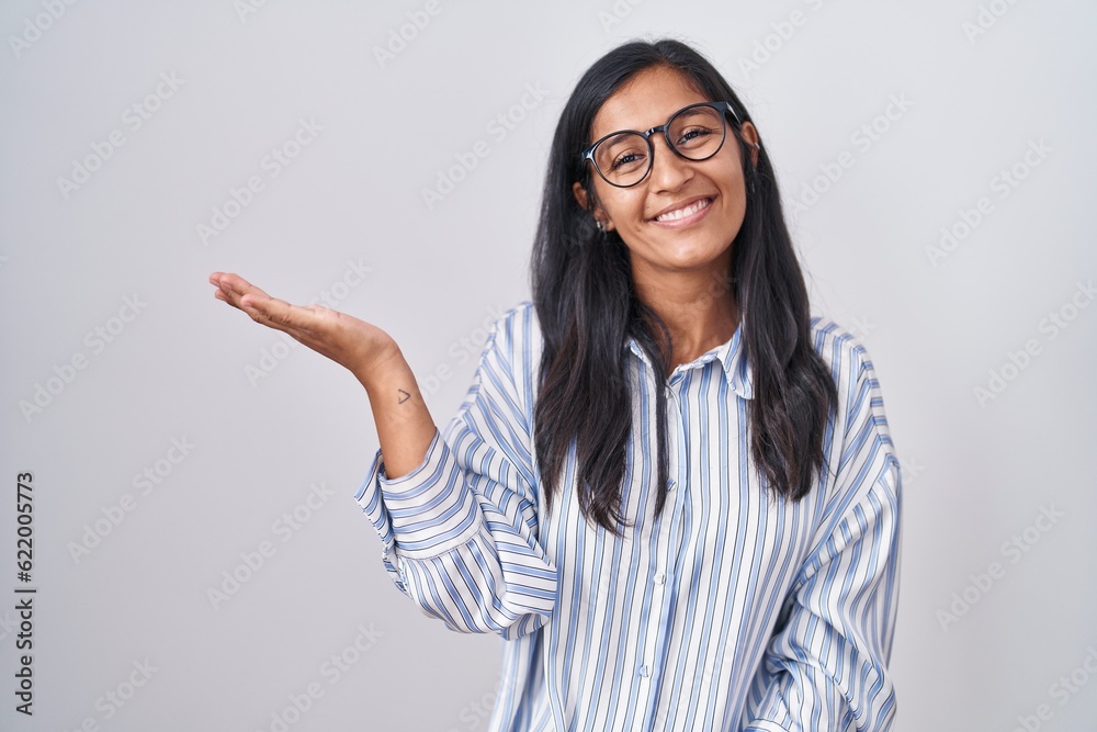 Young hispanic woman wearing glasses smiling cheerful presenting and pointing with palm of hand looking at the camera. Young hispanic woman wearing glasses smiling cheerful presenting and pointing with palm of hand looking at the camera.