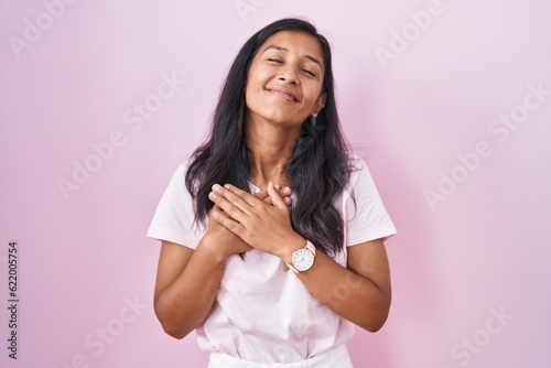 Young hispanic woman standing over pink background smiling with hands on chest with closed eyes and grateful gesture on face. health concept.