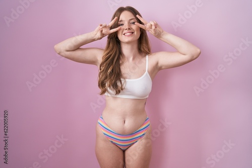 Caucasian woman wearing lingerie over pink background doing peace symbol with fingers over face, smiling cheerful showing victory © Krakenimages.com