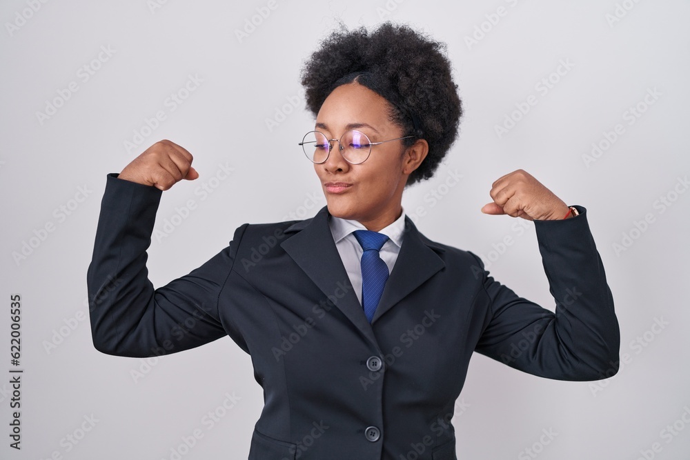 Beautiful african woman with curly hair wearing business jacket and glasses showing arms muscles smiling proud. fitness concept.