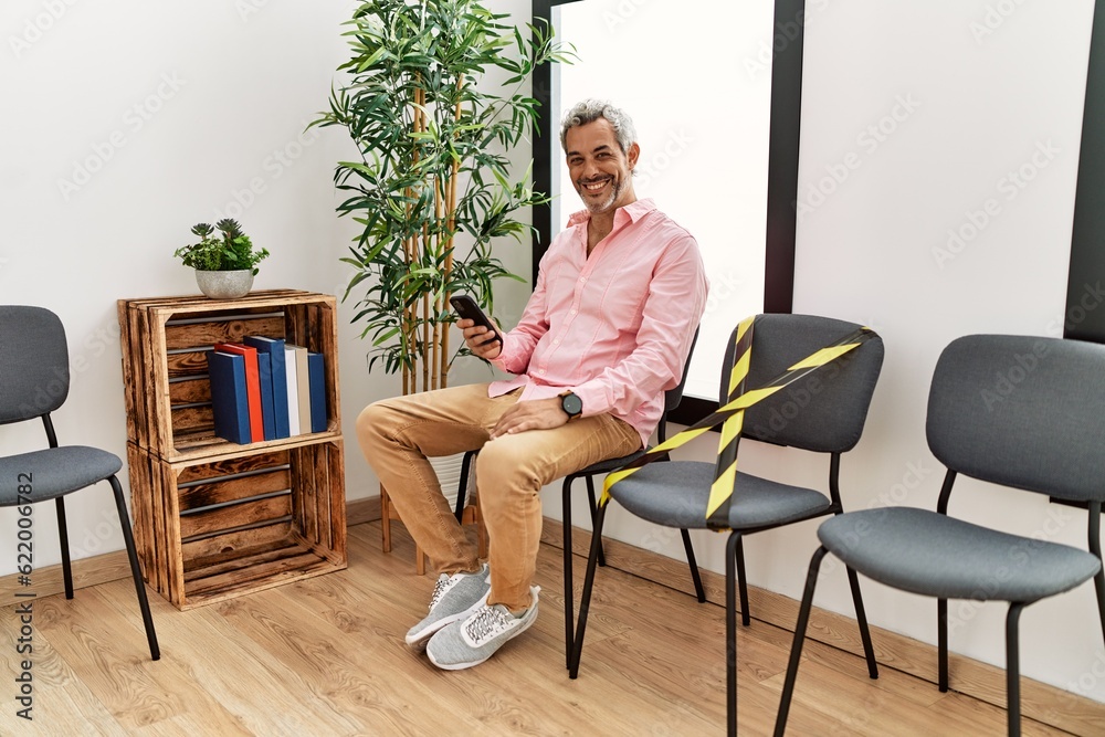 Middle age grey-haired man using smartphone sitting on chair at waiting room Middle age grey-haired man using smartphone sitting on chair at waiting room