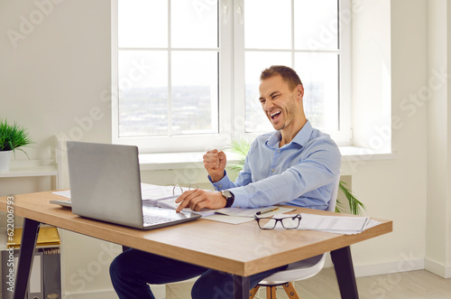Happy business man celebrating success in office. Young man sitting at working desk, looking at laptop computer, doing fist gesture, and saying Yes, I did it. Success, victory, satisfaction concept