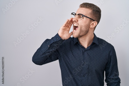 Young caucasian man standing over isolated background shouting and screaming loud to side with hand on mouth. communication concept.