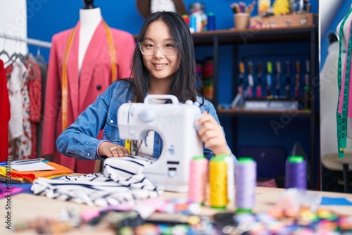 Young chinese woman tailor smiling confident using sewing machine at clothing factory