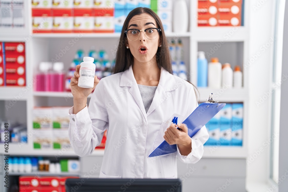 Young brunette woman working at pharmacy drugstore holding pills afraid and shocked with surprise and amazed expression, fear and excited face. Young brunette woman working at pharmacy drugstore holding pills afraid and shocked with surprise and amazed expression, fear and excited face.