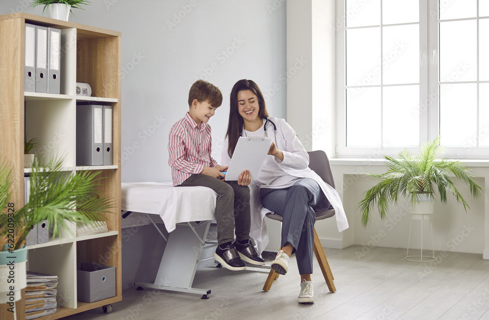 Portrait of a friendly smiling female child doctor pediatrician showing report file with appointment to a little boy patient sitting on the couch during medical examination in clinic. Portrait of a friendly smiling female child doctor pediatrician showing report file with appointment to a little boy patient sitting on the couch during medical examination in clinic.