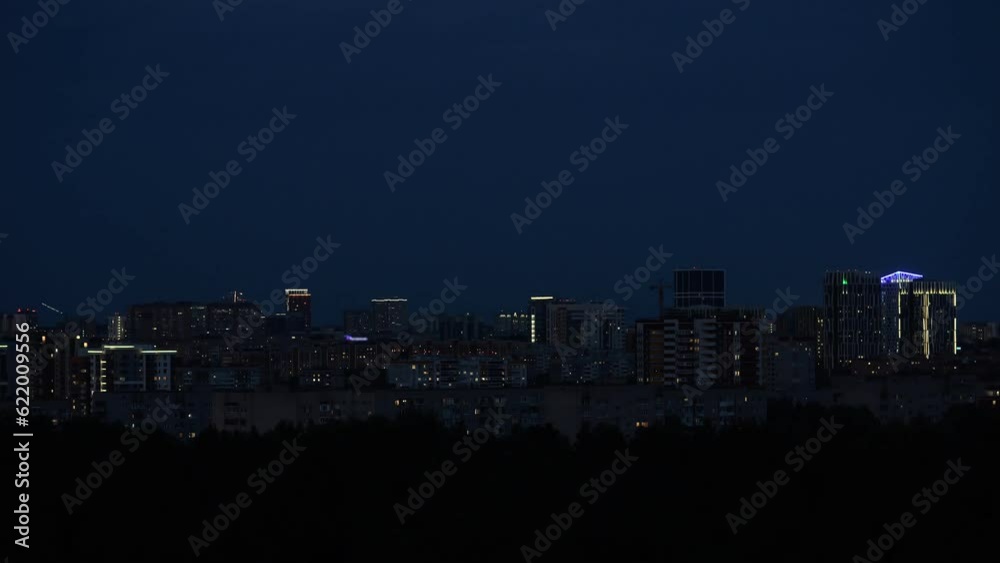 Transition of time from day to night of modern city near small area with trees. Concept of urban life. Buildings illuminated in evening time lapse
