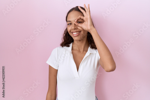 Young hispanic woman wearing casual white t shirt doing ok gesture with hand smiling, eye looking through fingers with happy face.