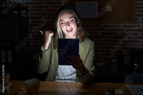 Blonde caucasian woman working at the office at night pointing to the back behind with hand and thumbs up, smiling confident