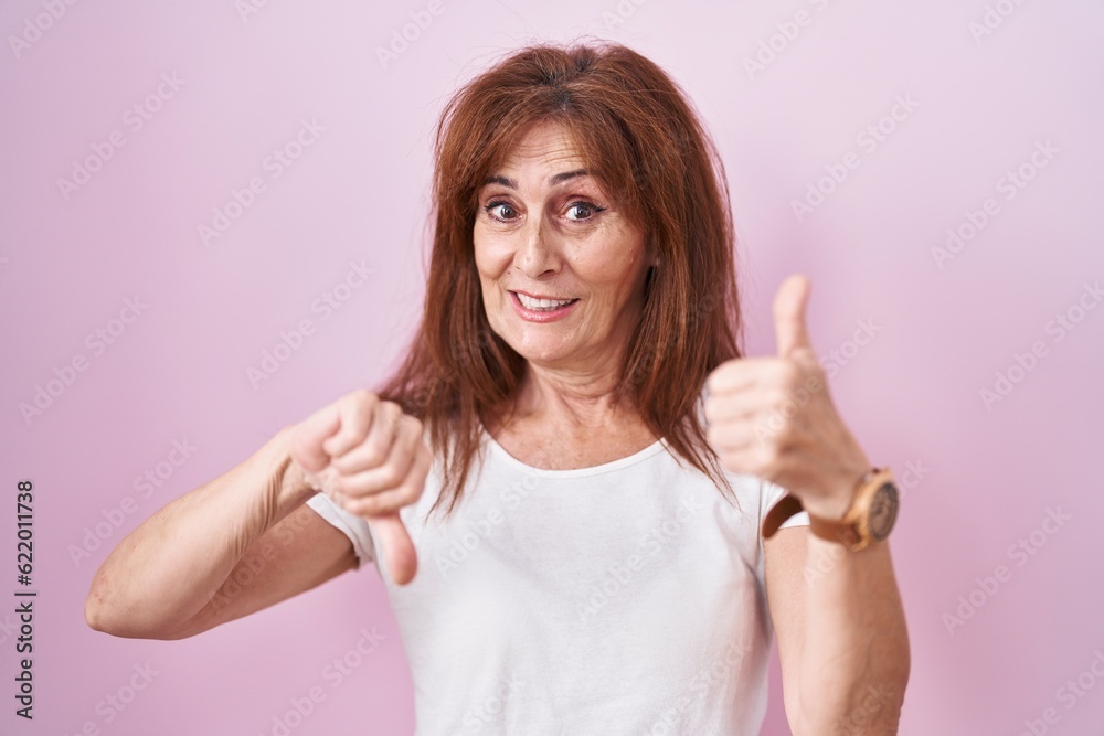 Middle age woman standing over pink background doing thumbs up and down, disagreement and agreement expression. crazy conflict Middle age woman standing over pink background doing thumbs up and down, disagreement and agreement expression. crazy conflict