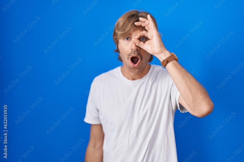 Middle age man standing over blue background doing ok gesture shocked with surprised face, eye looking through fingers. unbelieving expression. Middle age man standing over blue background doing ok gesture shocked with surprised face, eye looking through fingers. unbelieving expression.