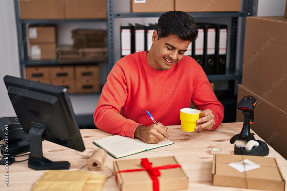 Young hispanic man ecommerce business worker writing on notebook drinking coffee at office Young hispanic man ecommerce business worker writing on notebook drinking coffee at office