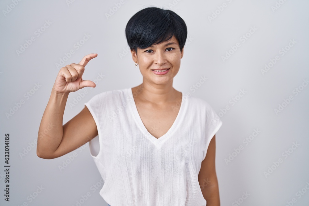 Young asian woman with short hair standing over isolated background smiling and confident gesturing with hand doing small size sign with fingers looking and the camera. measure concept. Young asian woman with short hair standing over isolated background smiling and confident gesturing with hand doing small size sign with fingers looking and the camera. measure concept.