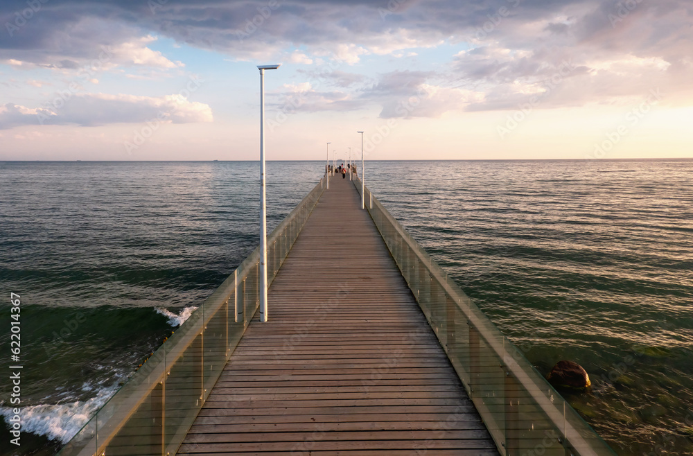 Perspective view of a long sea pier. There is a boardwalk and glass railings. Morning seascape. Human figures are visible in the distance. Background.