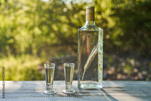 Bottle of crystal clear, famous and delicious Polish vodka with macerated corn ear inside and two old vintage glasses , served outdoor in summer sunny day. Popular drink in eastern european countries.
