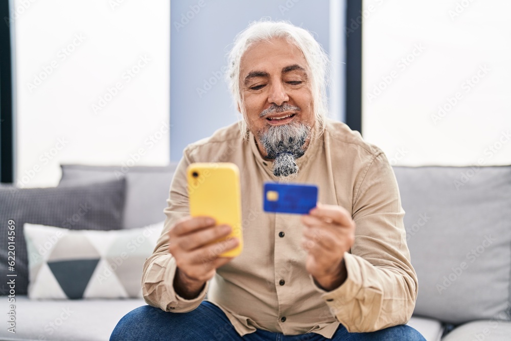Middle age grey-haired man using smartphone and credit card sitting on sofa at home Middle age grey-haired man using smartphone and credit card sitting on sofa at home