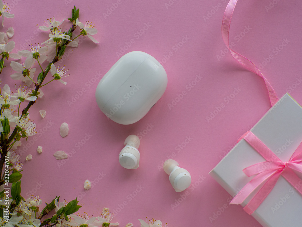 White wireless headphones on pink background with cherry flowers, gift box and copy space. Womens, Valentines and Mothers Day gift. Concept of music and elegance. Buying birthday gifts. 